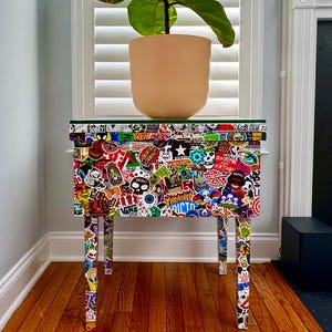 Op de afbeelding: Een kleine, rechthoekige tafel bedekt met kleurrijke stickers, met een glazen blad. De tafel heeft vier poten en staat voor een raam met een potplant erop. De plant heeft grote, groene bladeren en een beige pot.