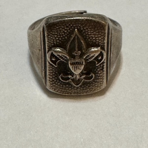 May include: A silver-toned ring with a rectangular face featuring a detailed fleur-de-lis emblem. The ring has a textured surface and a classic design, likely vintage or antique. The emblem includes a shield and a stylized lily.