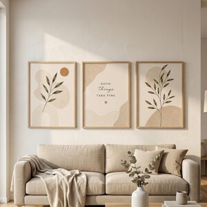 Op de afbeelding: Drie ingelijste wandkunstwerken met botanische illustraties en abstracte vormen in neutrale beige en bruine tinten. De middelste print bevat de tekst "GOOD THINGS TAKE TIME" met een klein hartje, terwijl de zijprints bladeren en takken tonen.