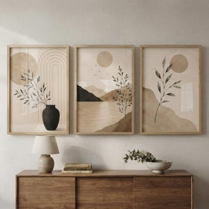Könnte beinhalten: Ein Set aus drei gerahmten Wandbildern mit beigen und sandfarbenen Landschaften, abstrakten Sonnen, einer schwarzen Vase mit einem Blattzweig und Bergsilhouetten. Die Bilder sind in hellen Holzrahmen über einem Sideboard platziert.