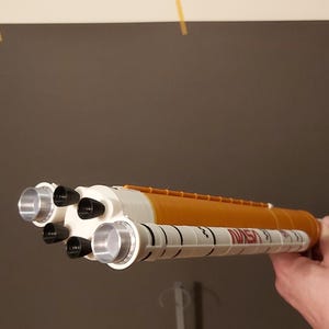 Op de afbeelding: Een speelgoedmodel van een NASA-spaceshuttle, wit en oranje van kleur, met zilveren motoruitlaten. Het NASA-logo staat op de zijkant. Het model wordt door een hand vastgehouden.