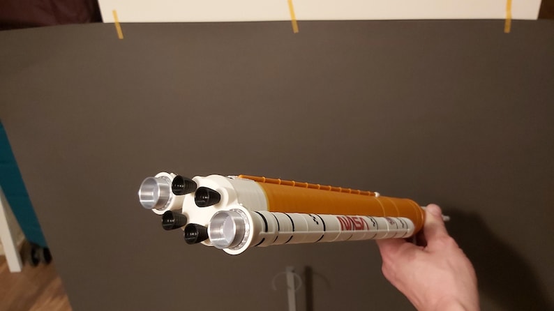 Op de afbeelding: Een speelgoedmodel van een NASA-spaceshuttle, wit en oranje van kleur, met zilveren motoruitlaten. Het NASA-logo staat op de zijkant. Het model wordt door een hand vastgehouden.