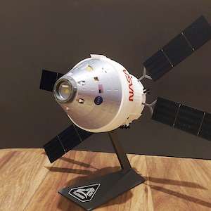Può includere: Un modello della navicella spaziale Artemis della NASA, con una capsula argentata e bianca e il logo NASA in rosso. Ha quattro pannelli solari neri ed è montato su un supporto nero con il logo Artemis. Il modello è esposto su una superficie di legno.