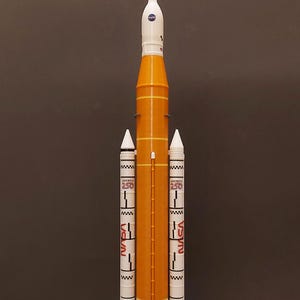 May include: A model rocket in orange and white, with the NASA logo and the word "America" in red. The model has two white side boosters and a base with the Artemis logo.