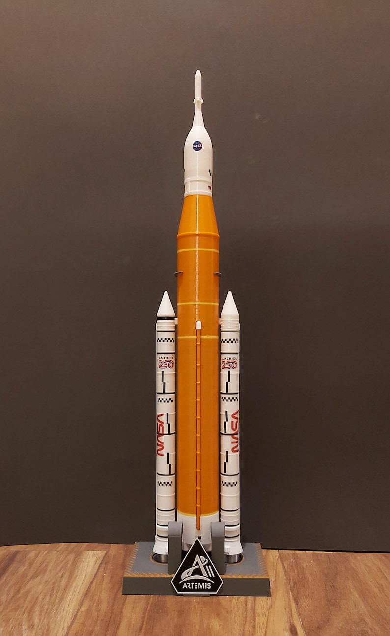 Op de afbeelding: Een modelraket in oranje en wit, met het NASA-logo en het woord "America" in het rood. Het model heeft twee witte zijboosters en een basis met het Artemis-logo.