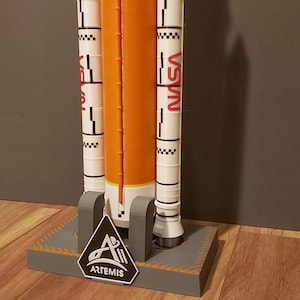 Op de afbeelding: Een modelraket met een oranje centraal lichaam en witte boosters, met het NASA-logo in rood. Het model is gemonteerd op een grijze basis met het Artemis-logo.