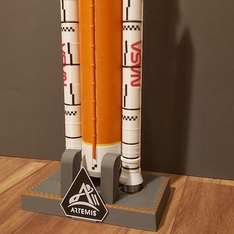 Op de afbeelding: Een modelraket met een oranje centraal lichaam en witte boosters, met het NASA-logo in rood. Het model is gemonteerd op een grijze basis met het Artemis-logo.