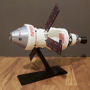 Può includere: Un modello bianco e argento di un'astronave con il logo NASA in rosso. Presenta pannelli solari e una bandiera americana. Il modello è esposto su un supporto nero, posto su una superficie di legno.