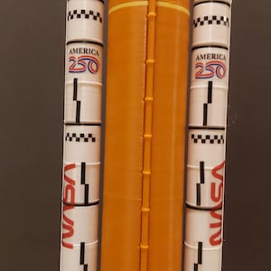 Op de afbeelding: Een modelraket met een oranje centraal lichaam en witte zijboosters. De boosters tonen de tekst "AMERICA 250" en "NASA" in het rood. De raket heeft een gedetailleerd ontwerp, inclusief zwart-witte dambordpatronen en een ladderachtige structuur.