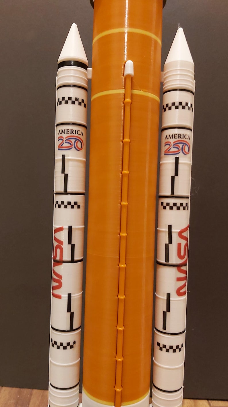 Op de afbeelding: Een modelraket met een oranje centraal lichaam en witte zijboosters. De boosters tonen de tekst "AMERICA 250" en "NASA" in het rood. De raket heeft een gedetailleerd ontwerp, inclusief zwart-witte dambordpatronen en een ladderachtige structuur.