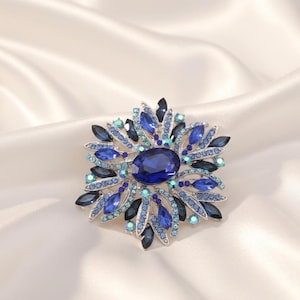 May include: A striking blue brooch with an oval central gem and petal-shaped stones. The brooch showcases various shades of blue and teal, forming a sparkling floral pattern. The jewellery is set against a white satin backdrop.