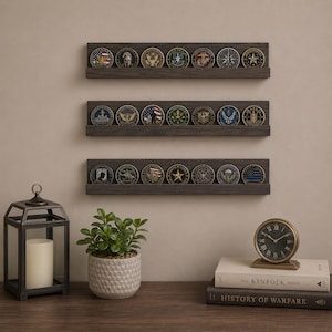 May include: Three dark gray wooden shelves displaying a collection of military challenge coins. The coins feature various emblems and designs. A decorative lantern, potted plant, and books with a clock are on the wooden surface below the shelves.