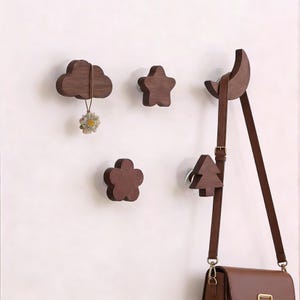 May include: Wooden wall hooks in cloud, star, flower, moon, and tree shapes. The hooks are dark brown and mounted on a white wall. A purse hangs from the moon hook, and a small flower decoration hangs from the cloud hook.