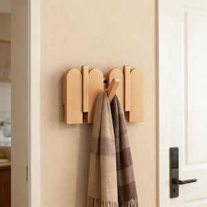 Könnte beinhalten: Ein wandmontierter Kleiderhaken aus Holz mit minimalistischem Design. Der Haken hat vertikale und bogenförmige Holzelemente. Ein beige-brauner Schal mit Karomuster hängt am zentralen Haken. Der Haken ist an einer hellbeigen Wand in der Nähe einer weißen Tür befestigt.