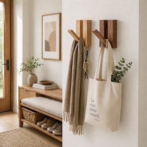 Op de afbeelding: Twee houten wandhaken, één licht en één donker, houden een sjaal, sleutels en een draagtas vast. De beige draagtas heeft de tekst "La nuit porte conseil". Op de achtergrond staat een bank met een kussen en schoenen.