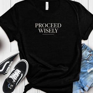 Op de afbeelding: Zwart T-shirt met de tekst "PROCEED WISELY" in een schreefletter. Het shirt wordt gecombineerd met zwart-witte sneakers en een spijkerbroek. Het shirt is in de taille geknoopt.