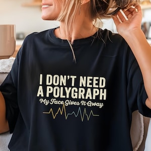May include: Black t-shirt with the text "I DON'T NEED A POLYGRAPH My Face Gives It Away" in a white, yellow, and blue design. The shirt features a heartbeat-like graphic below the text. The shirt is worn by a person.