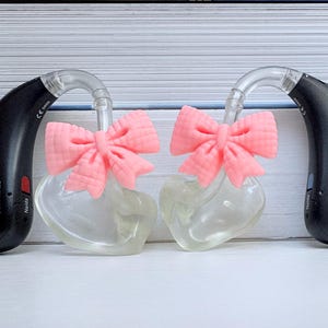 以下が含まれることがあります： ピンクのリボンがアクセントになった、黒いPhonak補聴器が2つ。透明なイヤモールドが付いています。補聴器は並んで配置され、デザインが際立っています。「Phonak Naida」の文字がデバイスに表示されています。