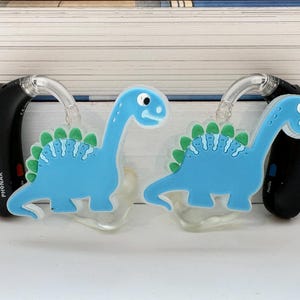 Puede incluir: Dos audífonos negros con tubos transparentes adornados con colgantes de dinosaurios azul claro. Los colgantes de dinosaurios tienen espinas verdes y detalles blancos. El fondo está borroso.