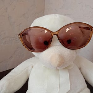 May include: Oversized sunglasses with a translucent amber-colored frame and gradient brown lenses. The frames feature a rounded, cat-eye shape with subtle horizontal ribbed detailing on the temples near the hinges, providing a classic look.