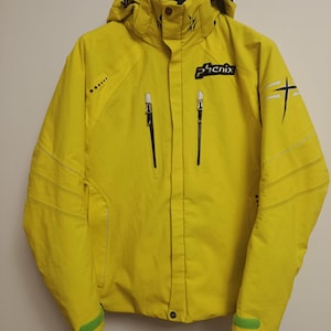 Puede incluir: Chaqueta de esquí amarilla brillante con capucha, cremalleras negras y la marca "phenix" en negro. La chaqueta tiene un diseño en blanco y negro en la manga derecha y detalles verdes en los puños.