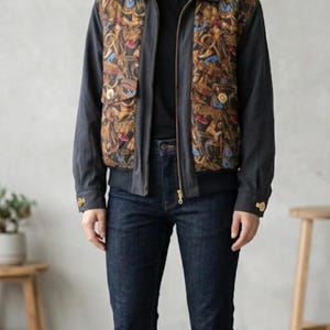May include: A dark gray jacket with a patterned front panel, paired with dark blue jeans. The jacket has a collar, a zipper, and gold-colored buttons. The jeans are a classic, straight-leg style.