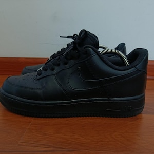 Pode incluir: Tênis Air Force 1 pretos. Os sapatos são totalmente pretos, incluindo os cadarços, a sola e o logotipo da Nike. Estes tênis de couro têm um design clássico de cano baixo. Estão sobre uma superfície de madeira.