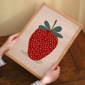 May include: A framed print featuring a stylized strawberry illustration. The strawberry is a vibrant red with white seed details and green leaves. The background is a light beige with small, evenly spaced dots. The frame is made of light-colored wood.