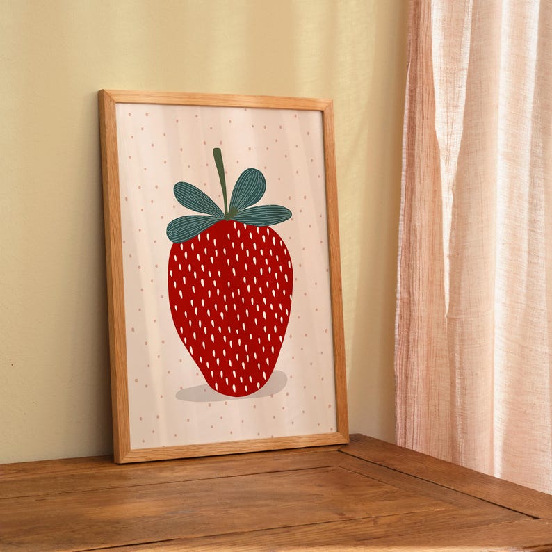 May include: A framed print featuring a stylized red strawberry with white seeds and green leaves. The artwork is set against a light pink background with small, evenly spaced dots. The frame is made of light-colored wood.