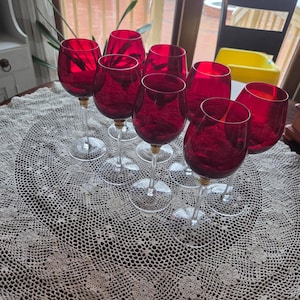 May include: A collection of eight ruby red wine glasses with clear stems and gold accents. The glasses are arranged on a round, cream-colored, crocheted lace tablecloth. The scene is set indoors with a view of a snowy outdoor scene.