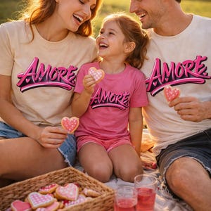 Peut inclure: Une famille porte des t-shirts assortis avec le mot "AMORE" en script rose et noir. Ils tiennent des biscuits en forme de cœur et profitent d'un pique-nique. Un panier en osier rempli de biscuits en forme de cœur et deux verres de liquide rose sont également visibles.