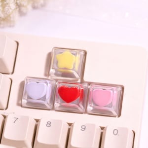 May include: A close-up of a computer keyboard with custom keycaps. The keycaps are clear with colorful shapes inside: a yellow star, a white heart, a red heart, and a pink heart. The keyboard keys are off-white.