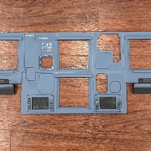 Puede incluir: Un panel rectangular gris claro con múltiples aberturas rectangulares, probablemente un componente de una estructura más grande. El panel tiene varias cajas rectangulares de color gris oscuro y dos formas octogonales negras en cada extremo. El panel está sobre una superficie de madera.