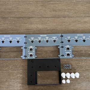 May include: A collection of aircraft cockpit panels and components. The panels are gray with white text and dials, labeled "SWITCHING". Additional black and white components and fasteners are included, arranged on a wooden surface.