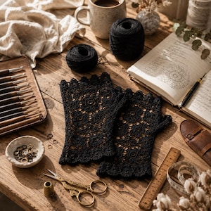 Puede incluir: Un par de guantes negros sin dedos con un intrincado patrón de ganchillo floral y bordes festoneados. Los guantes se muestran junto a un juego de ganchillos de madera, ovillos de lana negra, un cuaderno, tijeras, un dedal y una cinta métrica.