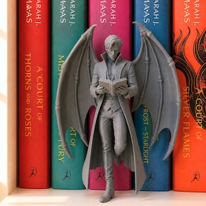 May include: A gray monochromatic figurine of a winged humanoid figure wearing a long coat and vest, holding an open book. The figure stands in front of five colorful book spines by Sarah J. Maas, including A Court of Thorns and Roses and Silver Flames.