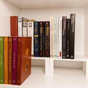 May include: A collection of books arranged on two white shelves. The left side features a series of colourful, uniform volumes titled Beltane, Ostara, Midsummer, Lughnasadh, Samhain, and Mabon. The right side holds various novels, including titles by J.R.R. Tolkien and Warcraft.
