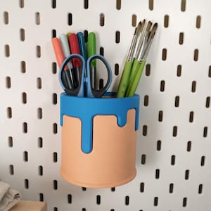 Puede incluir: Un organizador de escritorio cilíndrico de dos tonos, melocotón y azul, con un diseño de goteo, que contiene útiles de arte. El organizador contiene tijeras, pinceles y rotuladores de colores. El organizador está montado en una pared perforada blanca.