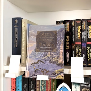 以下が含まれることがあります： 「Arda: Un viaje ilustrado por la Tierra Media y los reinos de Tolkien」というタイトルのハードカバー本。山のある沿岸都市の風景イラストが描かれており、様々なタイトルの本が並ぶ棚の中に置かれています。