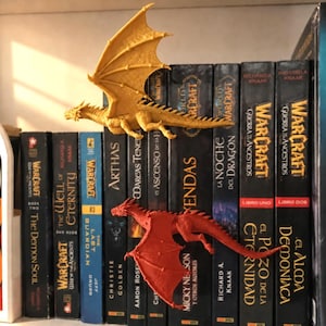 Pode incluir: Duas figuras de dragão de plástico, uma amarela e uma vermelha, estão posicionadas contra uma fileira de lombadas de livros de World of Warcraft. O dragão amarelo está acima do vermelho, ambos com asas, escamas e caudas longas.