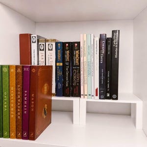 Può includere: Una collezione di libri disposti su due ripiani bianchi all'interno di un armadio bianco. I libri includono titoli come Mabon, Samhain, Lughnasadh, Midsummer, Ostara, The Last Guardian, The Demon Soul, The Well of Eternity e Historias de la Historia.