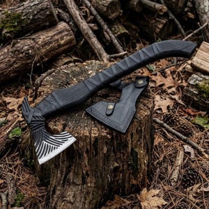May include: A black hand axe with a curved handle and a silver blade featuring intricate etched patterns. The axe head is partially covered by a black leather sheath secured with a brass snap button, resting on a textured wooden stump.