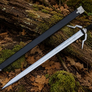May include: A silver and black sword with a black sheath rests on a bed of leaves and moss-covered wood. The sword has a silver blade, a black handle, and a decorative silver hilt. The sheath is black with silver detailing.