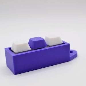 Könnte beinhalten: Ein lila Tastatur-Tastenhalter mit drei Tasten. Zwei weiße Tasten flankieren eine einzelne lila Taste. Der Halter ist rechteckig mit einer kleinen kreisförmigen Verlängerung. Der Hintergrund ist schlicht weiß.