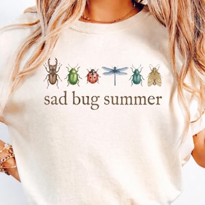 Könnte beinhalten: Ein cremefarbenes Kurzarm-T-Shirt mit einer horizontalen Reihe von sechs farbigen Insektenillustrationen, darunter ein Käfer, ein Marienkäfer und eine Libelle, mit dem braunen Schriftzug "sad bug summer" direkt unter den Insekten.