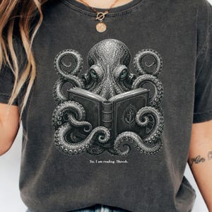May include: A charcoal grey t-shirt featuring a detailed graphic of an octopus with textured skin and blue eyes holding an open book with an anchor emblem on the cover. Below the illustration, the text reads: Sir, I am reading. Shoosh.