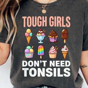 May include: A charcoal grey t-shirt featuring the text TOUGH GIRLS DON'T NEED TONSILS in white and peach lettering. The design includes eight colourful illustrations of various ice cream treats, including cones, sundaes, and popsicles in shades of pink, blue, brown, and yellow.