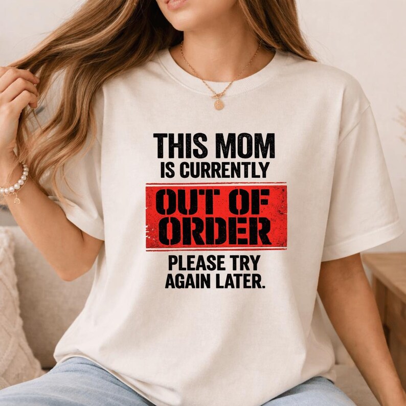Pode incluir: Camiseta creme com o texto "THIS MOM IS CURRENTLY OUT OF ORDER PLEASE TRY AGAIN LATER" em preto e vermelho. A camiseta &eacute; de estilo casual, adequada para o uso di&aacute;rio.
