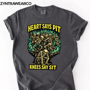 Puede incluir: Camiseta gris oscuro con un diseño gráfico de esqueletos y el texto "HEART SAYS PIT" y "KNEES SAY SIT" en amarillo. El diseño incluye llamas verdes y un esqueleto sentado en una silla.
