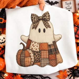 May include: A white sweatshirt featuring a graphic of a ghost with a leopard print bow and a patchwork-style body in shades of orange, brown, and tan. The ghost is positioned next to a matching patchwork pumpkin. The design has visible stitching.